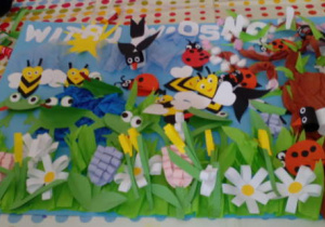 Obraz wykonanej przez dzieci pracy z napisem "Witaj Wiosno!". Przedstawia jaskółki, biedronki, pszczoły krążące nad trawą z pierwszymi kwiatami oraz stawem z żabkami. Z prawej strony mieści się zakwitające drzewo. W górze widać słoneczko.